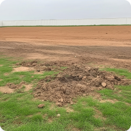 A cleared lot with a pile of excavated dirt in the foreground and a white fence in the back.